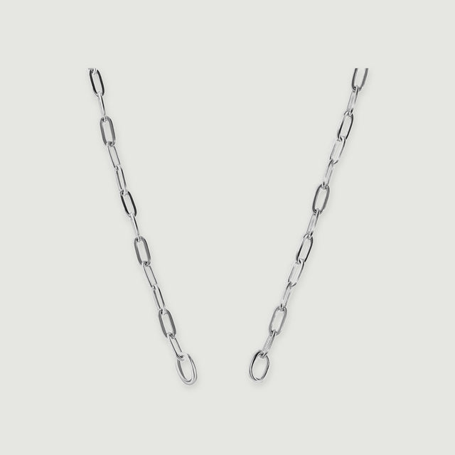 A polished silver chain with elongated links and two round end rings is centered on a pale grey surface.