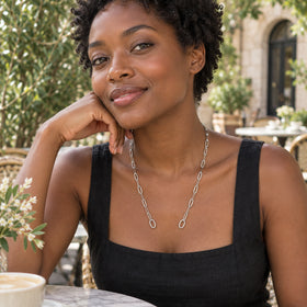 A woman in a black linen top wears a silver chain with two round end rings at an outdoor cafe.