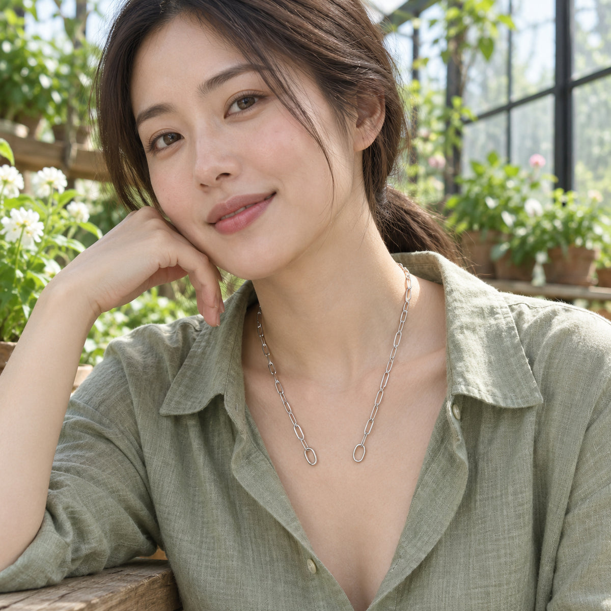 A woman in a sage green blouse wears an open silver chain beside greenhouse windows and plants.