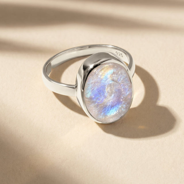 A silver moonstone ring rests on a pale textured tabletop with a shadow falling to the right.