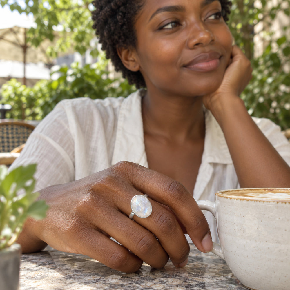A woman wears an oval silver moonstone ring while resting her hand beside a ceramic coffee cup outdoors.