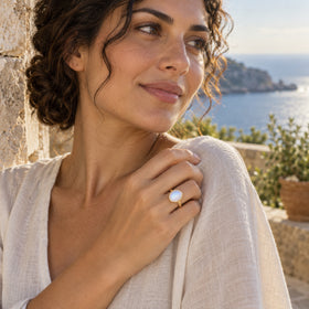 An oval moonstone ring in yellow gold vermeil sits on a hand touching a linen sleeve above the sea.