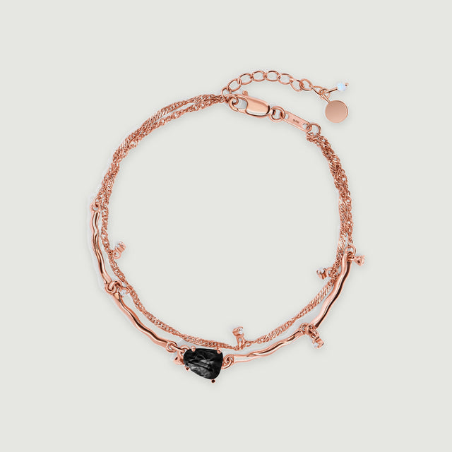 A rose gold bracelet with a black prong-set stone and small clear accents lies centered on a pale gray surface.