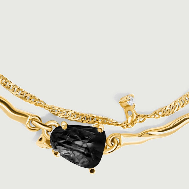 The yellow gold links and fine chain frame a rough black stone with small clear stones nearby.