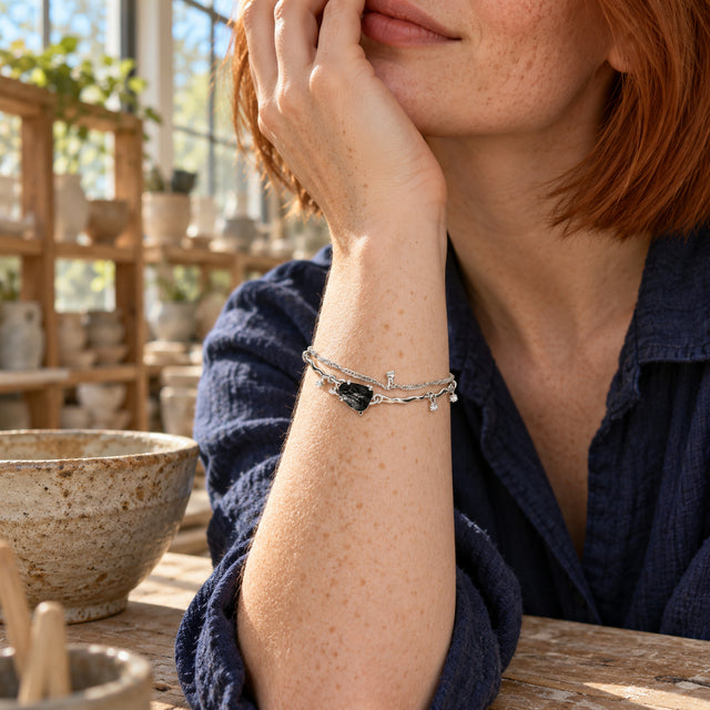 A freckled woman wears the silver black-stone bracelet on a raised wrist beside a ceramic bowl in a sunlit studio.