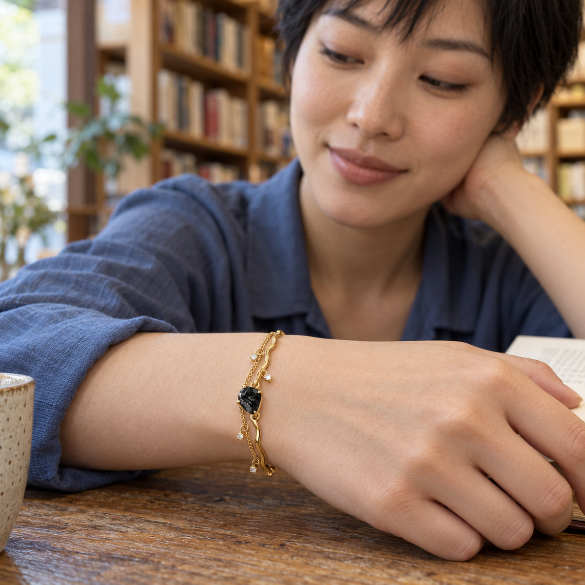 A yellow gold bracelet with a rough black stone rests around a wrist above a wooden reading table in a bookshop.