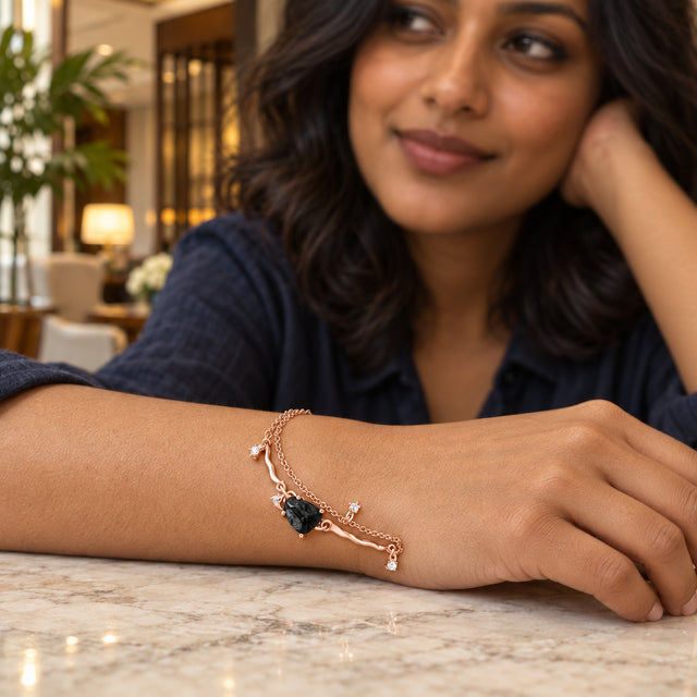 A rose gold bracelet with a dark textured stone is worn on a relaxed wrist resting on a pale stone table in a lounge.