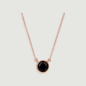 A rose gold necklace with a single round black pendant is centered against a light gray surface.