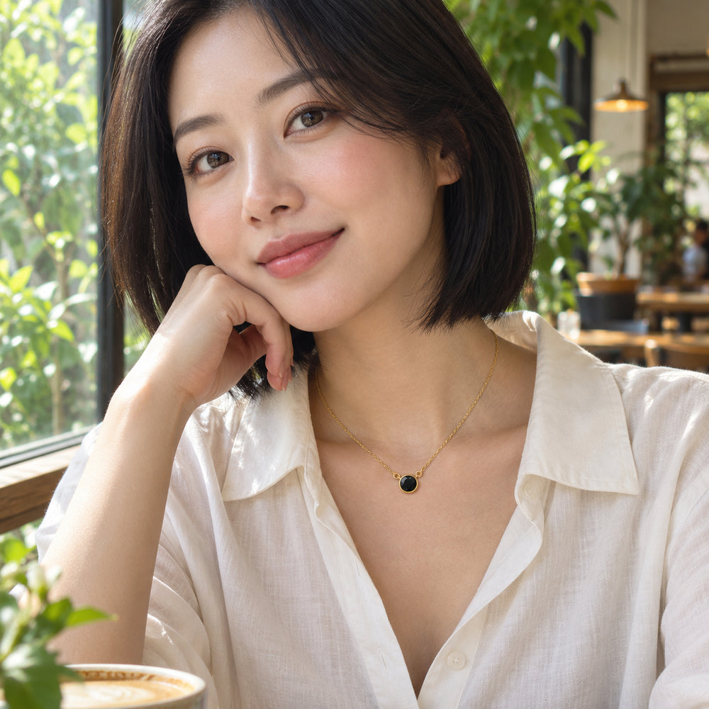 A woman in a white blouse wears the yellow gold black onyx necklace beside a leafy cafe window.