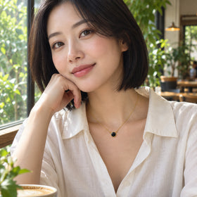 A woman in a white blouse wears the yellow gold black onyx necklace beside a leafy cafe window.