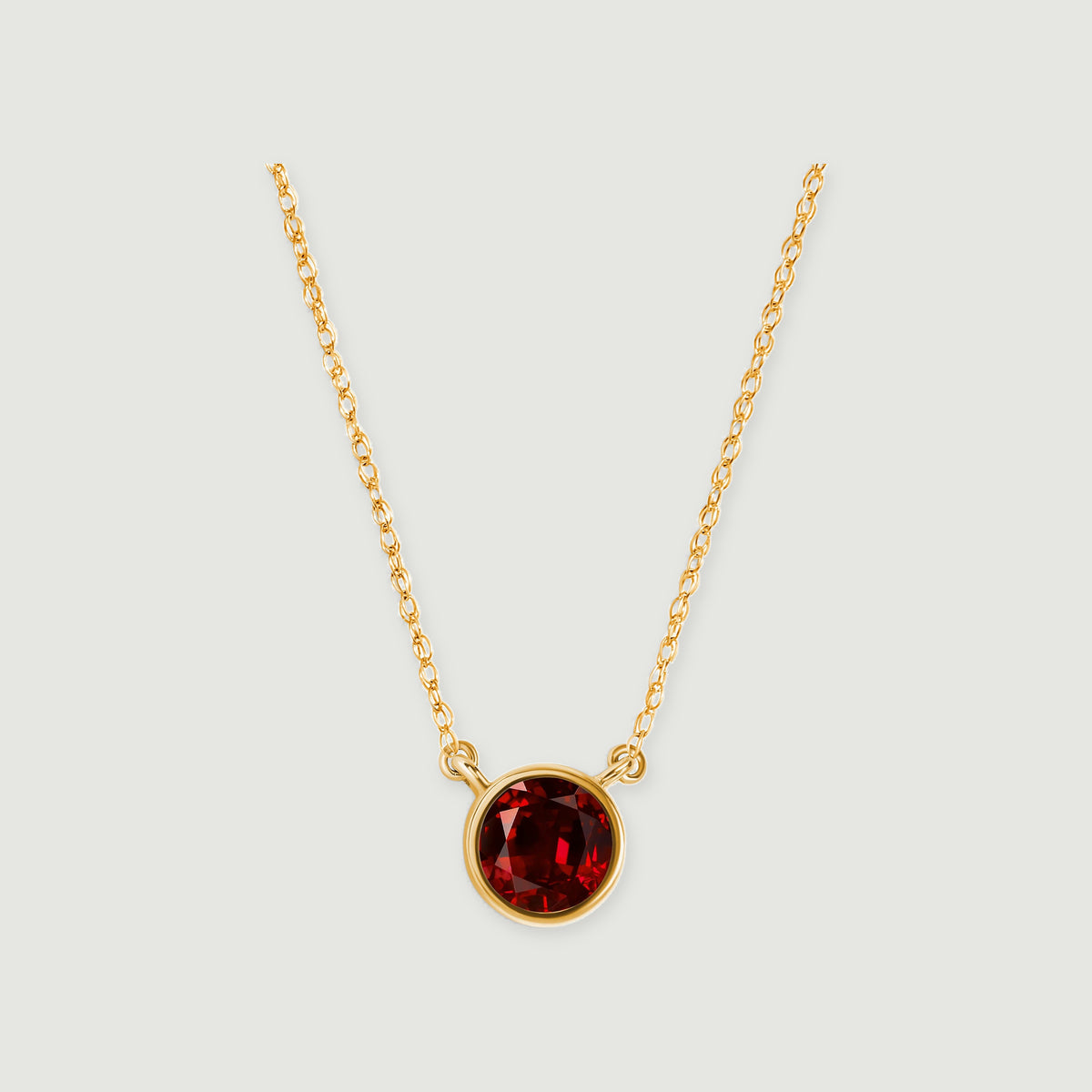 A round red garnet in a yellow gold bezel hangs between two lengths of fine chain on a light gray background.