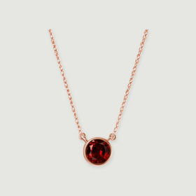 A polished rose gold bezel surrounds a faceted red garnet, with the chain rising on both sides.