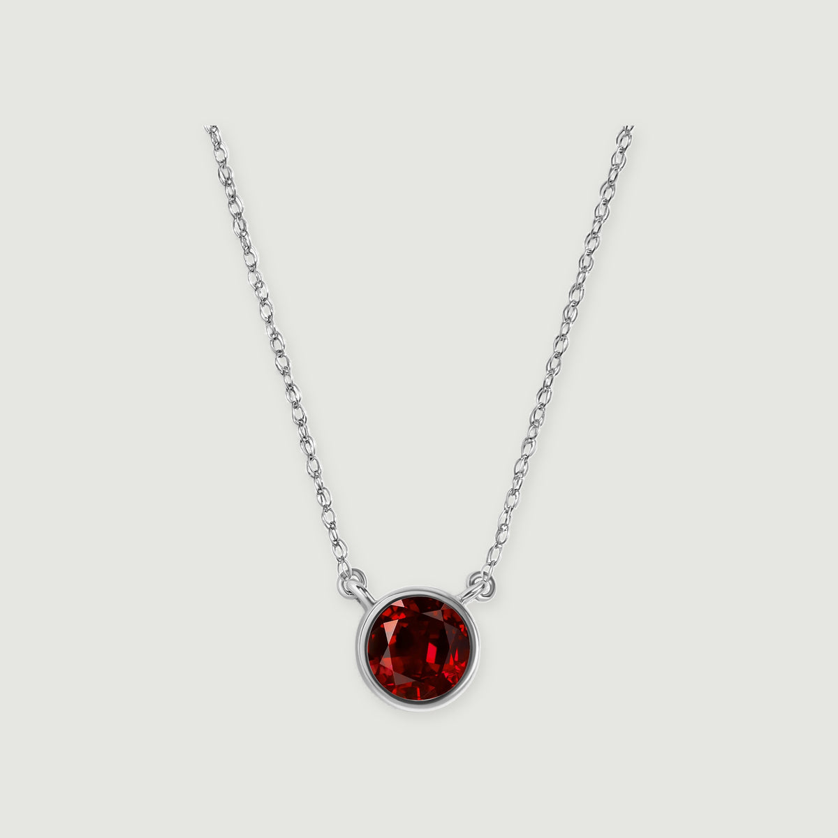 A white gold necklace displays a deep red round garnet against a softly shaded light gray surface.