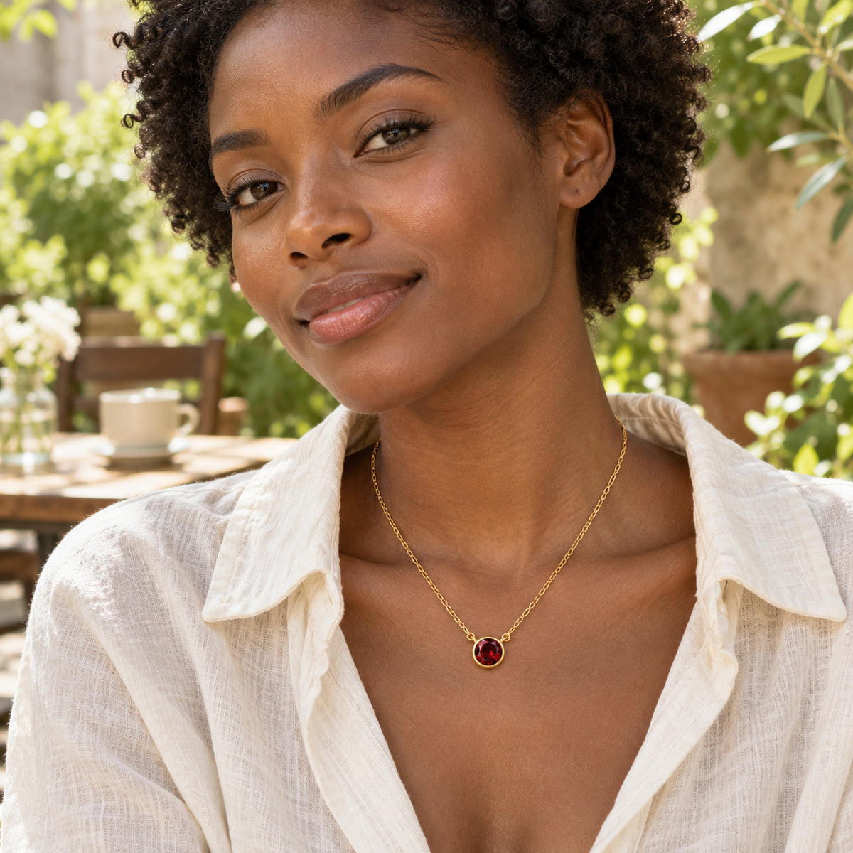 A woman with short curly hair wears the yellow gold garnet necklace with an ivory linen blouse in a leafy cafe courtyard.