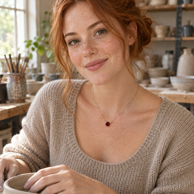 A red-haired woman wears the rose gold garnet necklace above an oat-colored sweater while holding a ceramic bowl in a pottery studio.