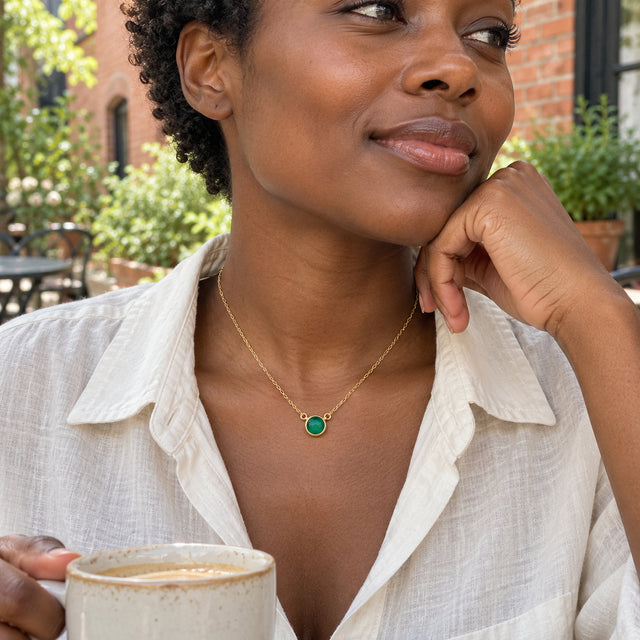 A woman in a white linen shirt wears a yellow gold green onyx necklace while seated with a cup in a brick courtyard.