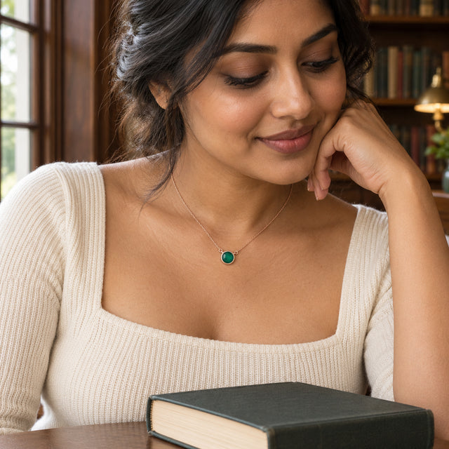 A woman in a cream square-neck top wears a rose gold green onyx necklace beside a library window and a closed book.