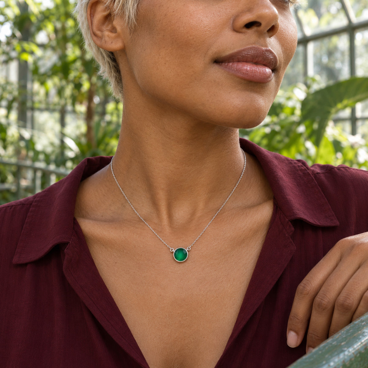 A woman with short pale hair wears a white gold green onyx necklace above a burgundy blouse in a leafy greenhouse.