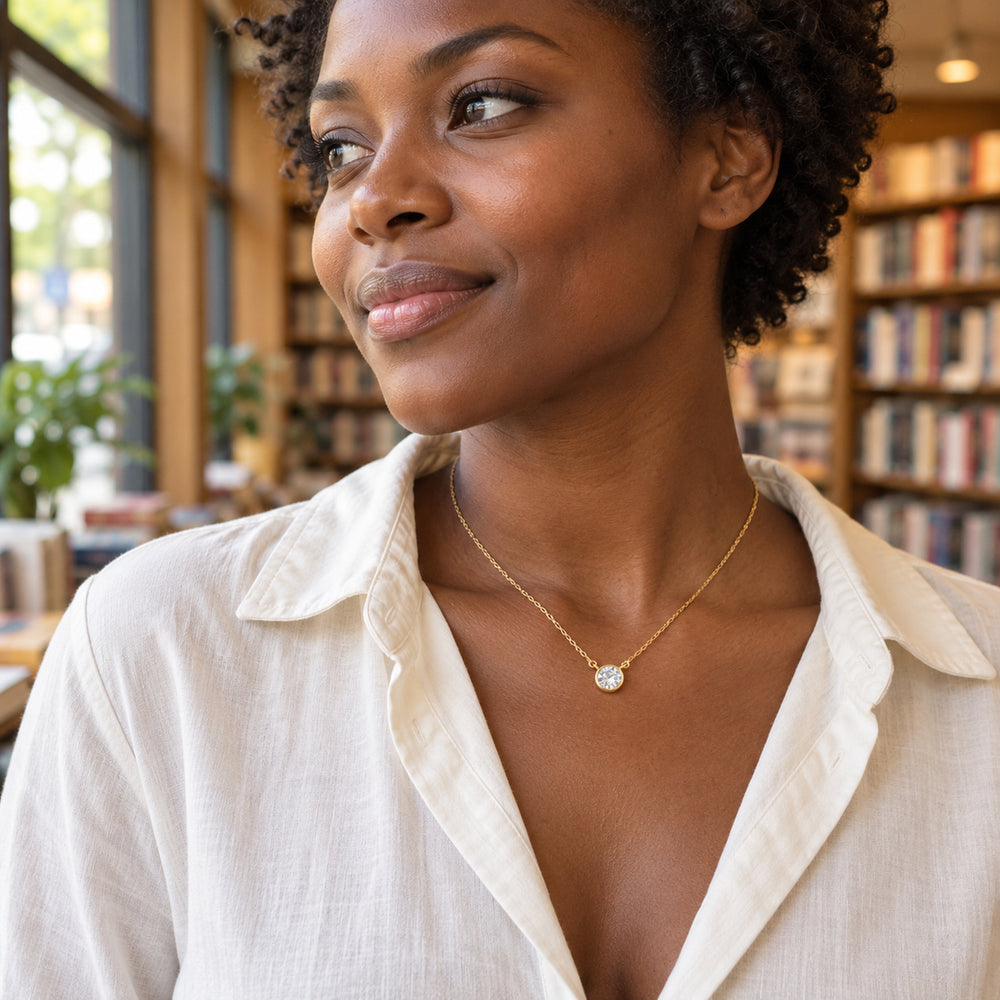 A woman in an ivory shirt wears a yellow gold white topaz necklace beside a bookstore window.