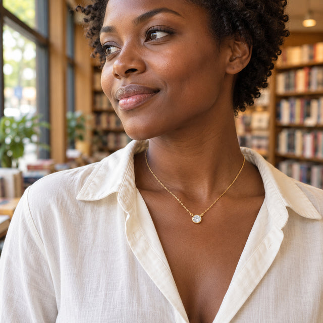 A woman in an ivory shirt wears a yellow gold white topaz necklace beside a bookstore window.