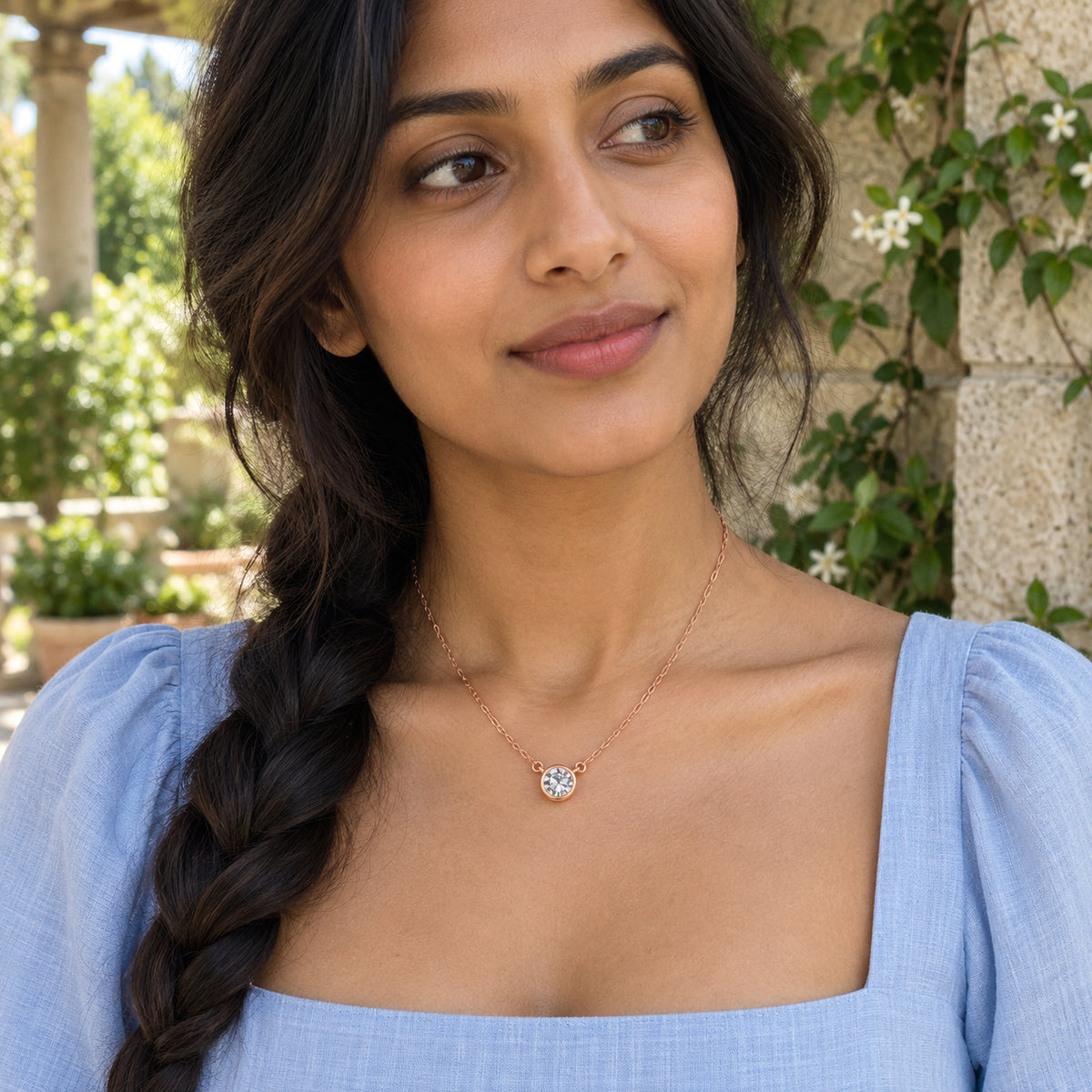 A rose gold white topaz necklace rests above a pale blue neckline on a woman with braided hair in a garden.