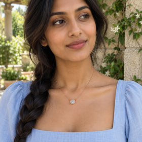 A rose gold white topaz necklace rests above a pale blue neckline on a woman with braided hair in a garden.