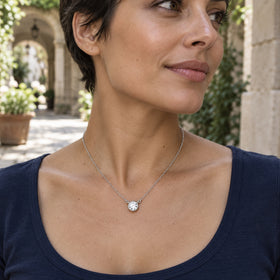 A woman in a navy scoop-neck top wears a white gold circular topaz pendant in a stone courtyard.