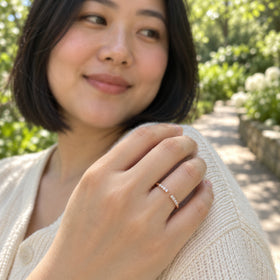 The rose gold diamond ring is shown on a hand resting against a cream knit cardigan in a leafy garden.