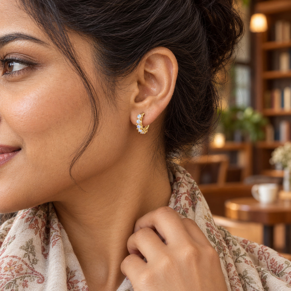 A yellow gold moonstone hoop is shown on an ear beneath dark swept-back hair in a warm book cafe.
