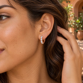 A rose gold moonstone hoop is worn with brown hair brushed behind the ear in a leafy terracotta courtyard.