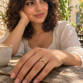 A yellow gold ring with three clear stones is shown on a hand resting on a wooden cafe table.