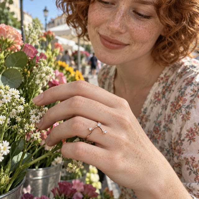 A rose gold wave ring with three clear stones is worn on a freckled hand beside fresh flowers.