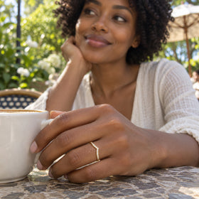 A yellow gold diamond ring is worn on a hand beside a white coffee cup at an outdoor cafe.
