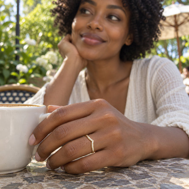 A yellow gold diamond ring is worn on a hand beside a white coffee cup at an outdoor cafe.