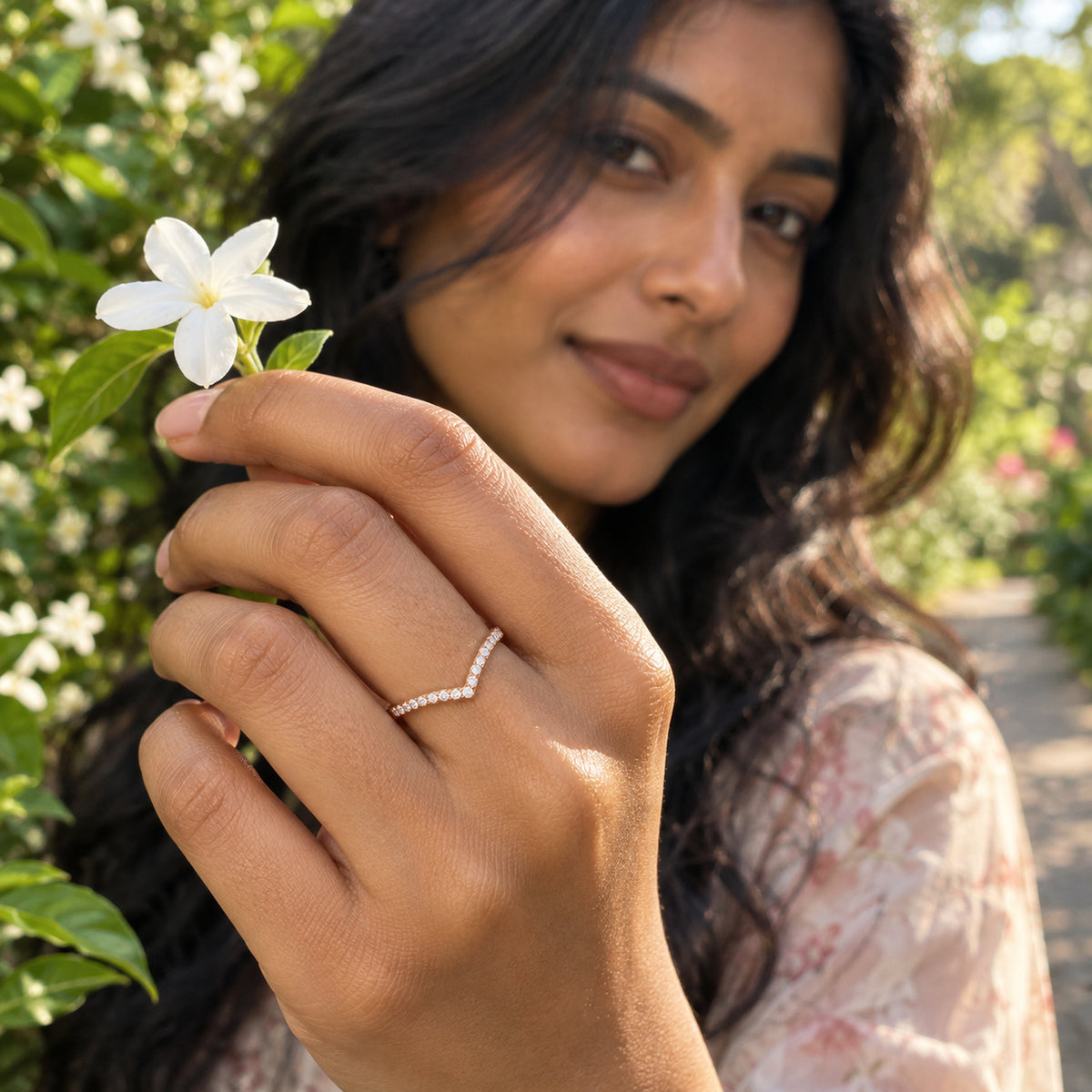 A rose gold diamond band sits on a finger as a woman holds a small white flower in a garden.