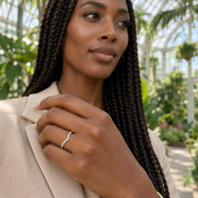 A white gold diamond ring is shown on a hand against a beige jacket with leafy plants behind it.