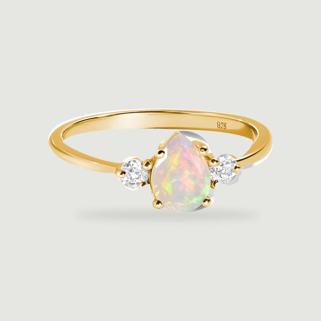 A yellow gold ring with a pear-shaped opal and two round clear stones rests on a light gray surface.