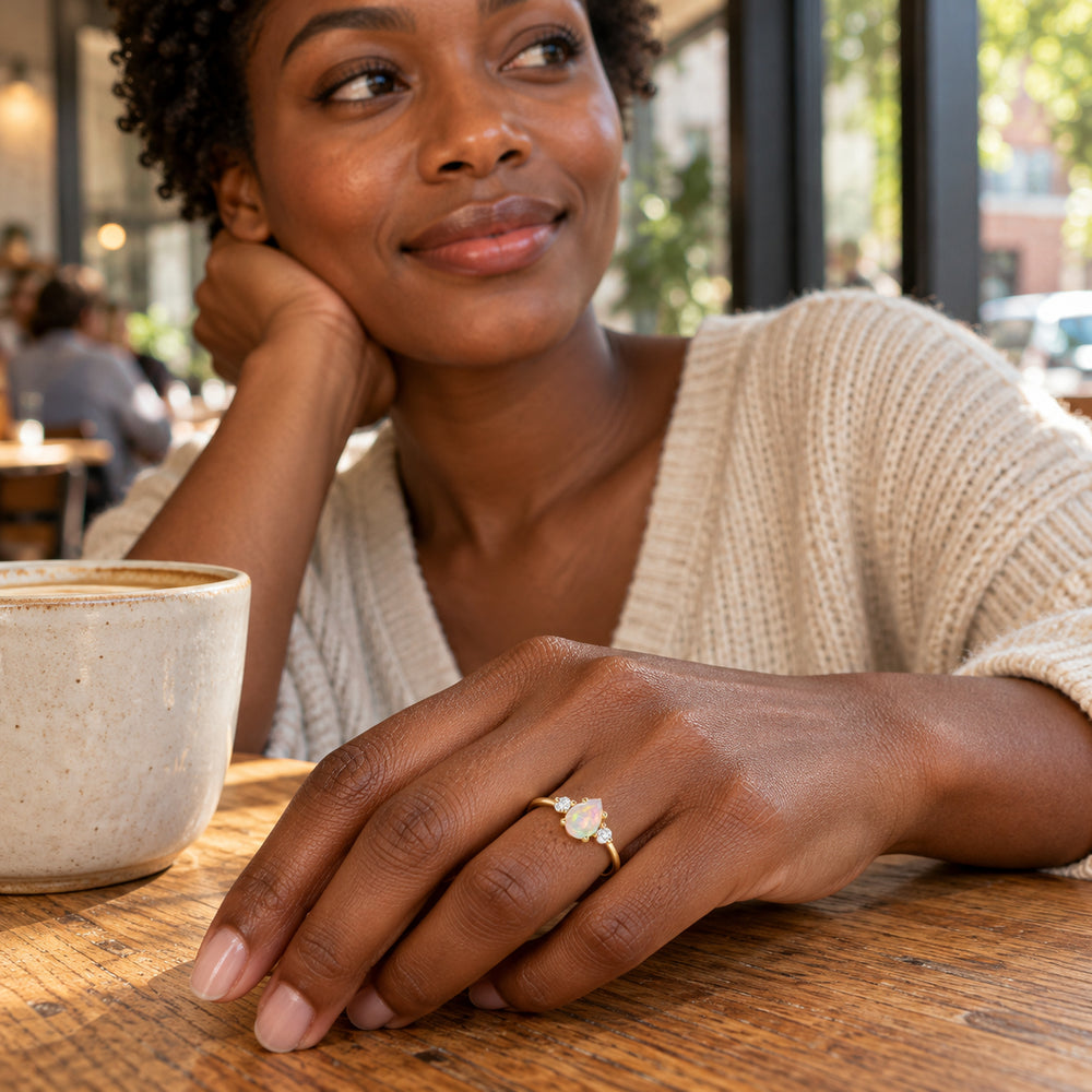 A woman wears a yellow gold opal ring with two clear side stones while resting her hand beside a ceramic cup on a wooden cafe table.