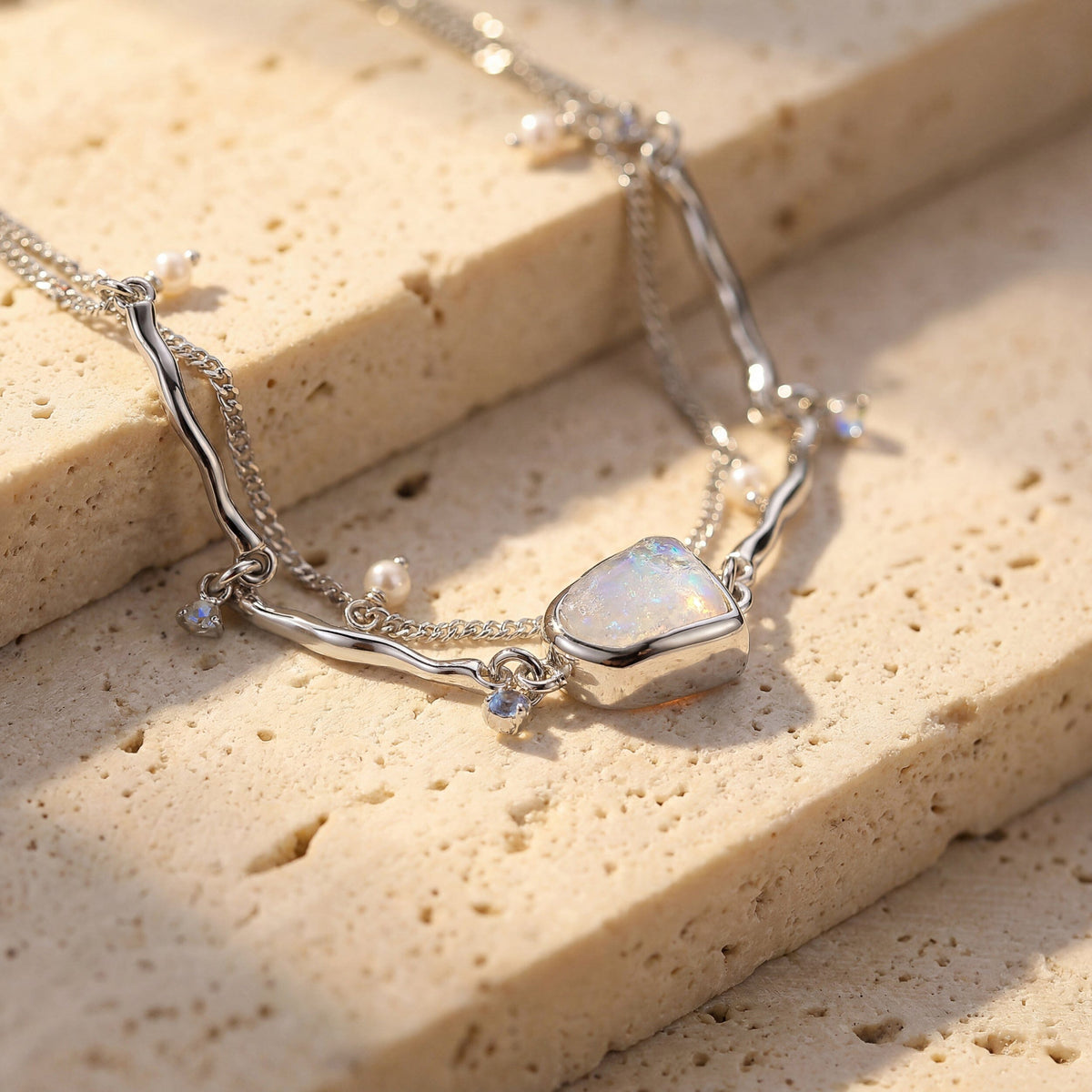 The silver moonstone bracelet drapes across pale stone steps with a pearl and blue charm beside the bezel.