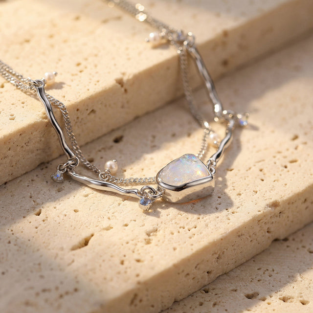 The silver moonstone bracelet drapes across pale stone steps with a pearl and blue charm beside the bezel.