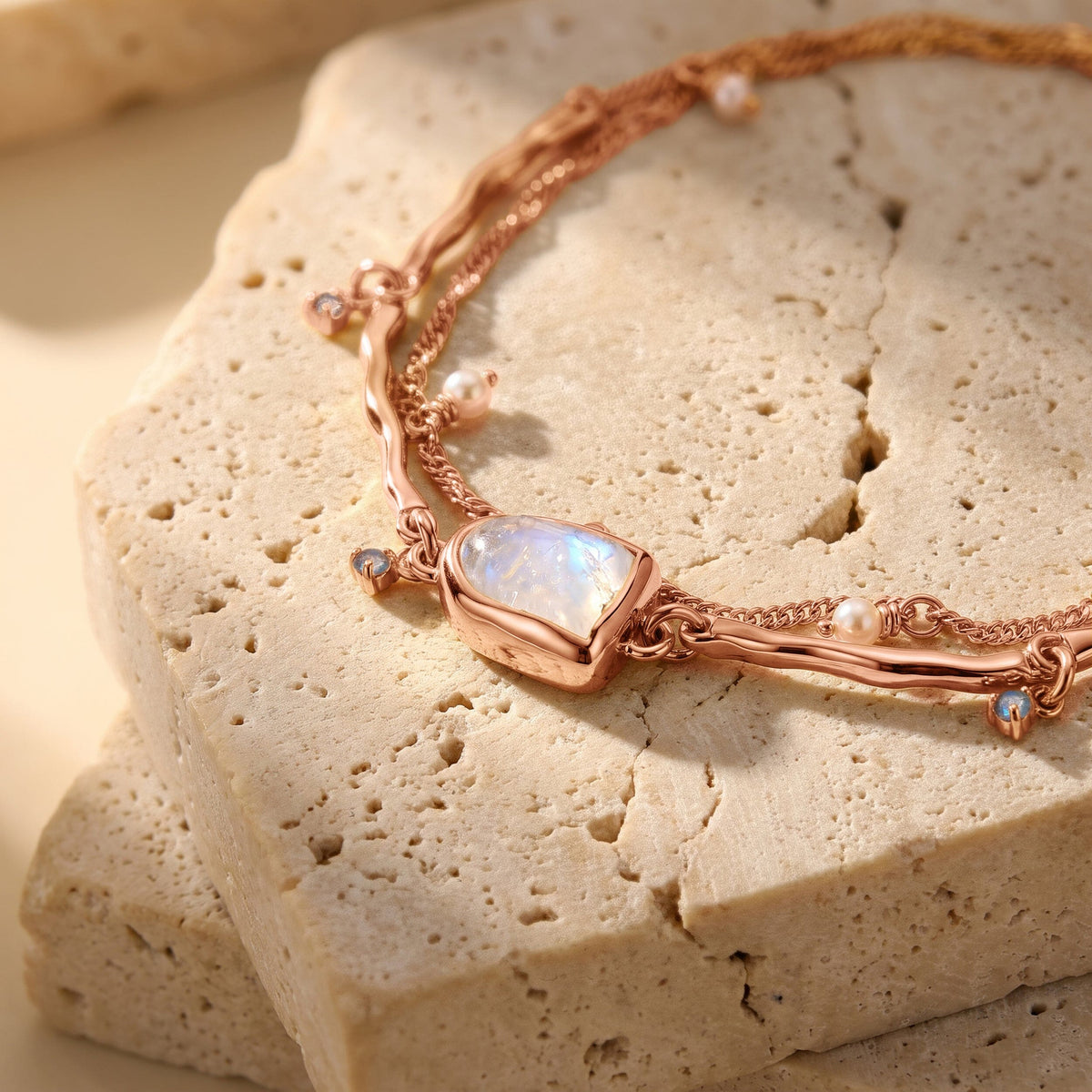A rose gold moonstone bezel sits on a stone slab beside rounded pearls and small blue dangling stones.