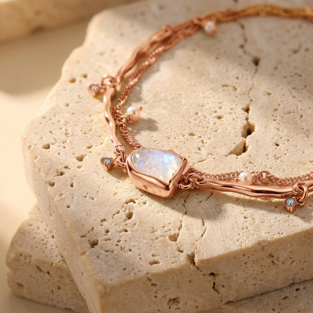 A rose gold moonstone bezel sits on a stone slab beside rounded pearls and small blue dangling stones.