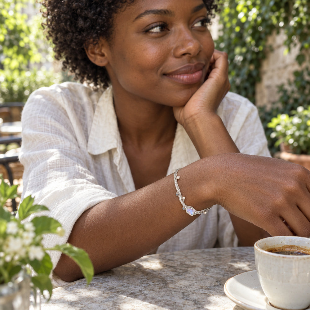 A woman wears the silver moonstone and pearl bracelet beside an espresso cup in a leafy courtyard.