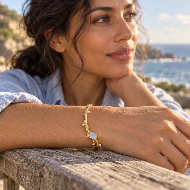 The yellow gold moonstone bracelet circles a wrist resting on a wooden railing with the sea behind.