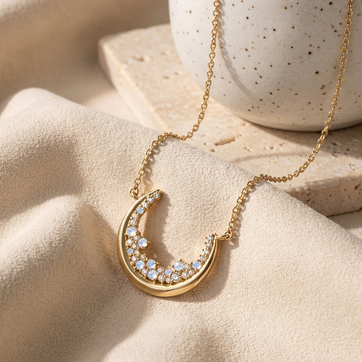 The gold crescent necklace rests across cream linen beside a speckled ceramic cup and pale stone.