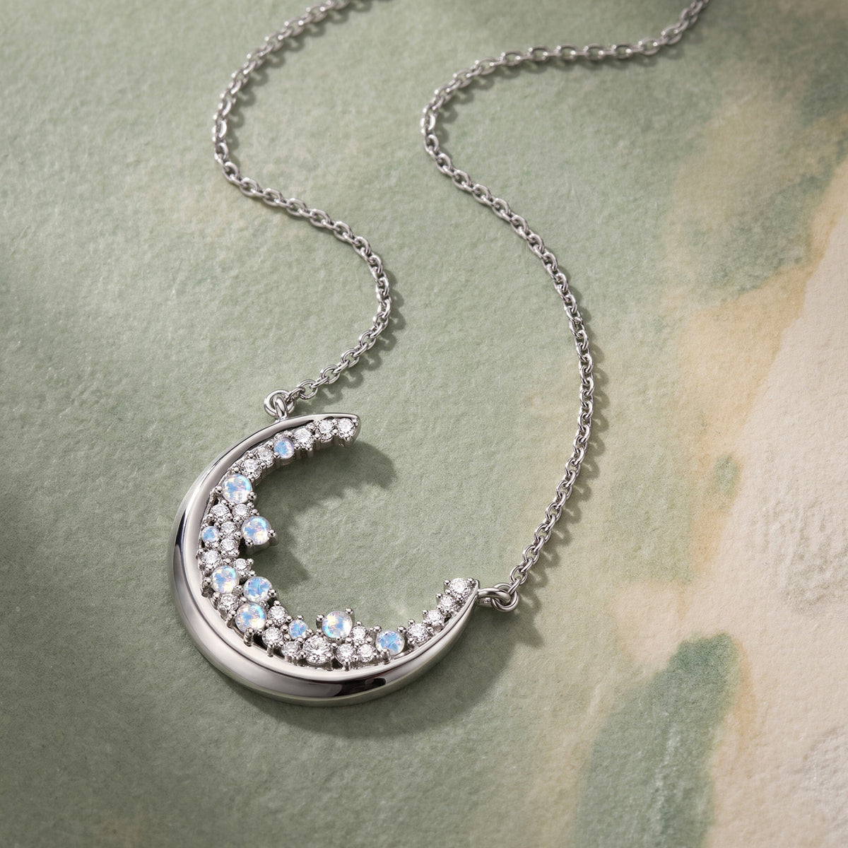 A silver crescent pendant and fine chain lie diagonally on a clean textured surface in soft daylight.