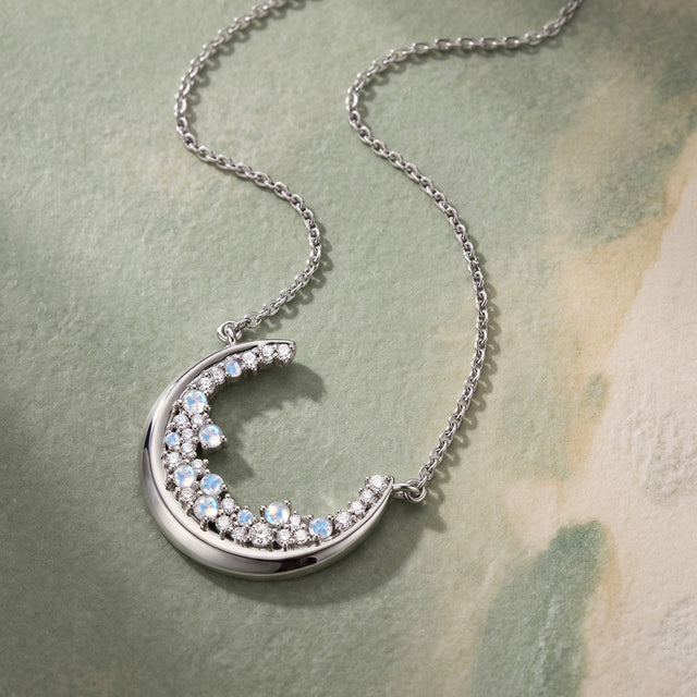 A silver crescent pendant and fine chain lie diagonally on a clean textured surface in soft daylight.