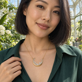 A woman in a green blouse wears the gold crescent necklace among sunlit plants in a glass conservatory.