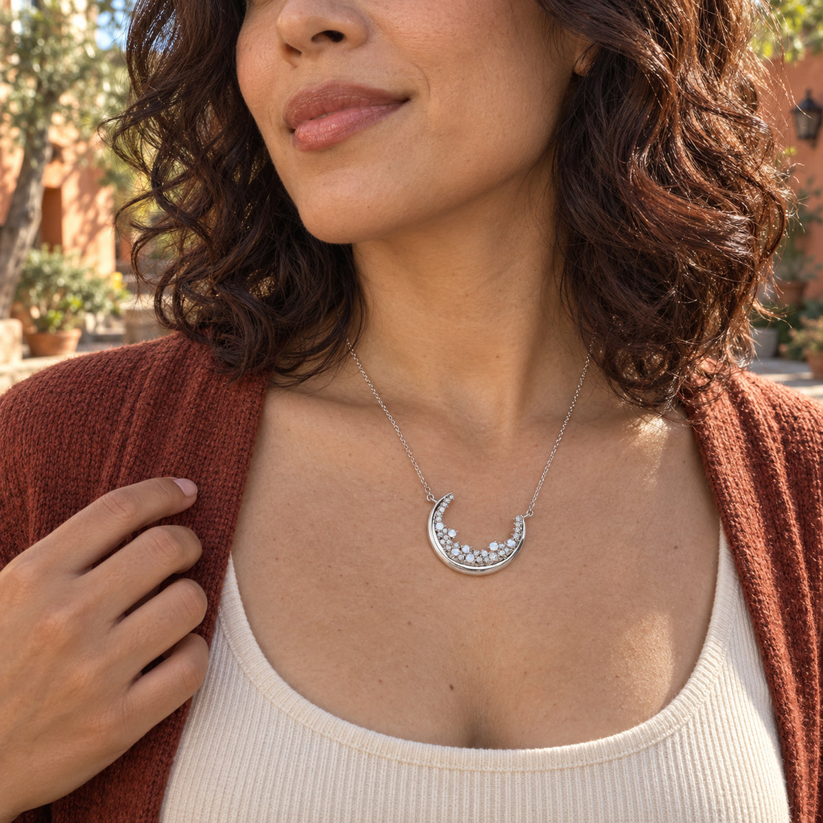 A woman with dark curls wears the silver crescent necklace above a cream top and rust cardigan in a sunny courtyard.