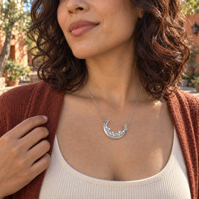 A woman with dark curls wears the silver crescent necklace above a cream top and rust cardigan in a sunny courtyard.